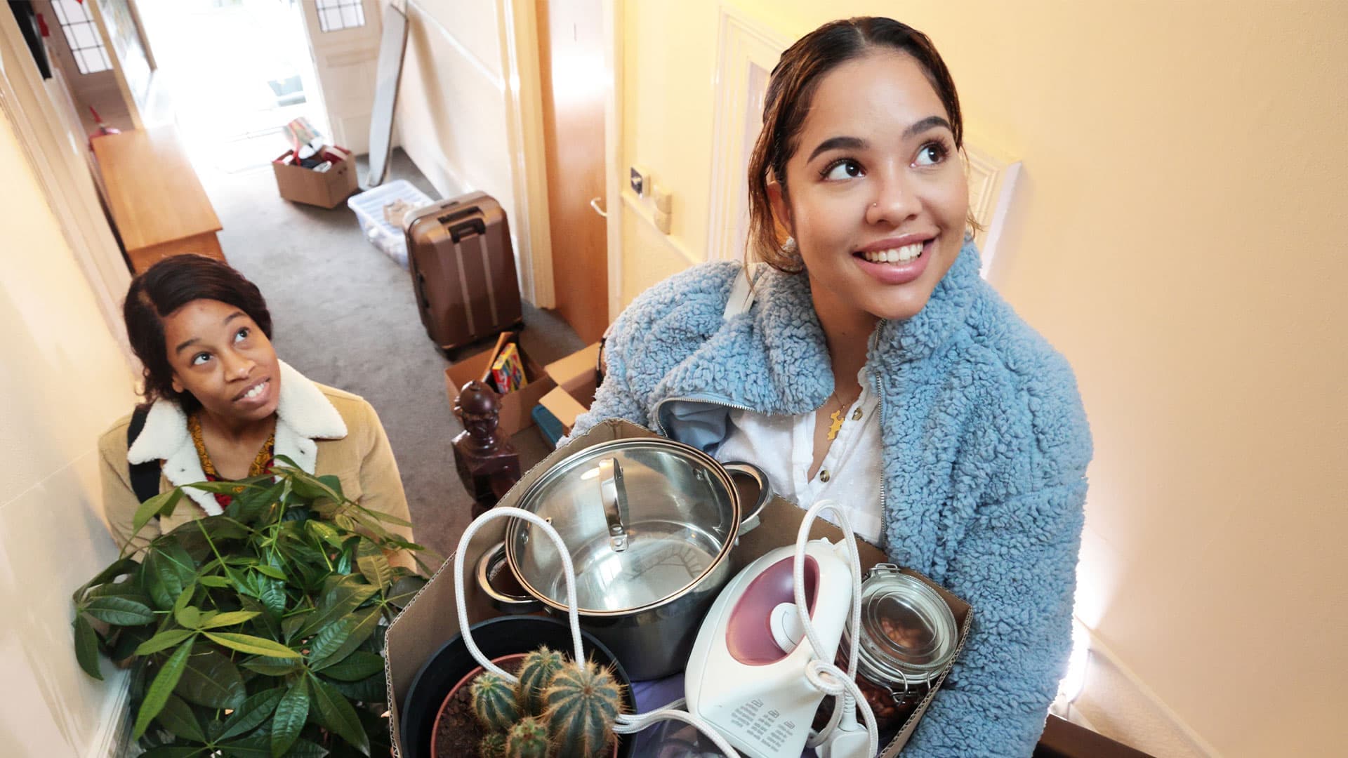 A person carries a box filled with household items, including a pot, iron, and plant, while navigating a well-lit hallway.
